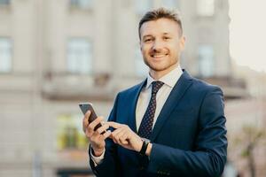 Satisfied businessman holds smart phone, reads email from investor, happy to recieve good news about business company, stands in urban territory, wears black suit and elegant tie. Technology concept photo