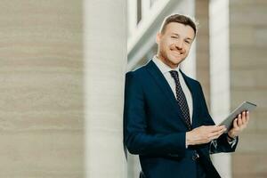 Portrait of stylish male entrepreneur has cheerful expression, holds digital tablet, reads financial news on web page, uses free internet connection, wears formal suit and tie. Career concept photo