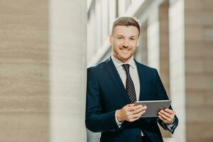 Confident cheerful male lawyer reads business news, has gentle smile, dressed in formal clothes, poses in urban setting. Businessman checks email or updates profile on digital tablet computer photo