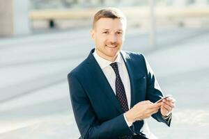 imagen al aire libre de un exitoso empresario masculino, usa traje negro formal, camisa blanca con corbata, sostiene un teléfono inteligente y revisa la notificación en la calle, envía un mensaje de texto o revisa el buzón de correo electrónico en línea foto