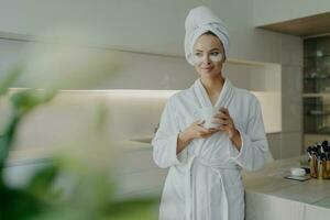 Relaxed woman in bathrobe, hair wrapped, tea in hand, rests with under-eye patches, post-spa, modern kitchen ambiance. photo