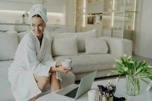 Relaxed woman in bathrobe with tea or coffee, sits on sofa, looks aside, works on laptop after shower. photo