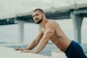 Motivated bearded man performs push-ups, exerting effort on a bridge, gazes into the distance. Athletic, bare-chested, working out outdoors. photo