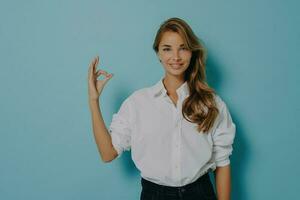 Confident female model in white shirt and jeans, shows okay gesture, assures quality, isolated on blue background. photo