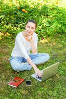 mujer joven sentada en el césped verde en el parque de la ciudad trabajando en una computadora portátil. concepto de negocio independiente foto