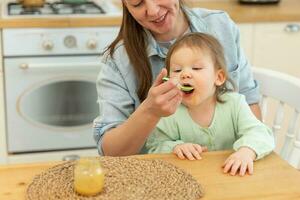 Happy family at home. Mother feeding her baby girl from spoon in kitchen. Little toddler child with messy funny face eats healthy food at home. Young woman mom giving food to kid daughter. photo