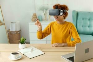 African american young woman wearing using virtual reality metaverse VR glasses headset at home. Girl touching air during VR experience on virtual reality helmet. Simulation hi-tech videogame concept. photo