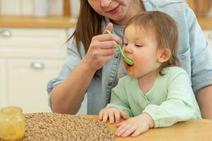 Happy family at home. Mother feeding her baby girl from spoon in kitchen. Little toddler child with messy funny face eats healthy food at home. Young woman mom giving food to kid daughter. photo