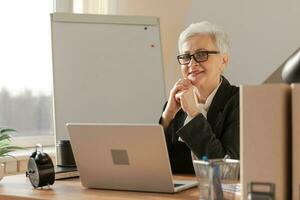 Portrait of confident stylish european middle aged senior woman at workplace. Stylish older mature 60s gray haired lady businesswoman sitting in modern office. Boss leader teacher professional worker. photo