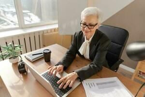 Confident stylish european middle aged senior woman using laptop at workplace. Stylish older mature 60s gray haired lady businesswoman sitting at office table. Boss leader teacher professional worker. photo