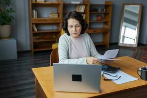 Middle aged senior woman sit with laptop and paper document, Pensive older mature lady reading paper bill pay online at home managing bank finances calculating taxes planning loan debt pension payment photo