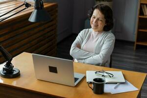 Confident stylish european middle aged senior woman using laptop at home. Stylish older mature 60s lady sitting at table looking at computer screen typing chatting reading writing email. photo