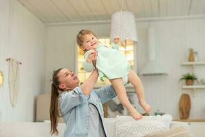 Happy family at home. Mother lifting in air little toddler child daughter. Mom and baby girl playing having fun together at home. Mother hugs baby with love care. Young mother kid rest in living room. photo