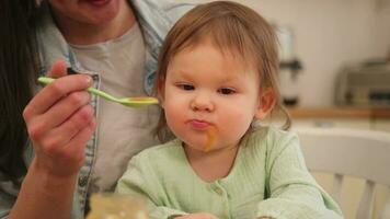 contento familia a hogar. madre alimentación su bebé niña desde cuchara en cocina. pequeño niñito niño con sucio gracioso cara come sano comida a hogar. joven mujer mamá dando comida a niño hija. foto