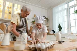 contento familia en cocina. abuela y nieta niño cocinar en cocina juntos. abuela enseñando niño niña rodar fuera masa hornear galletas. casa trabajo en equipo Ayudar familia generaciones concepto. foto