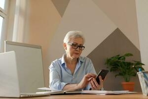 Confident stylish european middle aged senior woman using smartphone at workplace. Stylish older mature 60s gray haired lady businesswoman with cell phone in office. Boss leader using internet apps. photo