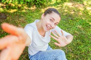 Young funny girl take selfie from hands with phone sitting on green grass park or garden background photo