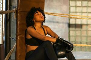 Woman fighter girl power. African american woman fighter with boxing gloves sitting on boxing ring waiting and resting after fight. Strong powerful girl in gym. Strength fit body workout training. photo