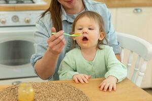 Happy family at home. Mother feeding her baby girl from spoon in kitchen. Little toddler child with messy funny face eats healthy food at home. Young woman mom giving food to kid daughter. photo