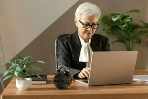 Confident stylish european middle aged senior woman using laptop at workplace. Stylish older mature 60s gray haired lady businesswoman sitting at office table. Boss leader teacher professional worker. photo