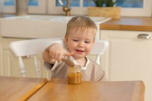 Happy family at home. Baby boy feeding himself in kitchen. Little boy with messy funny face eats healthy food. Child learns eat by himself holding spoon. Self feeding. photo