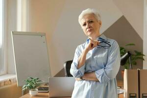 Portrait of confident stylish european middle aged senior woman at workplace. Stylish older mature 60s gray haired lady businesswoman in modern office. Boss leader teacher professional worker. photo