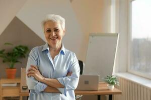 Portrait of confident stylish european middle aged senior woman at workplace. Stylish older mature 60s gray haired lady businesswoman in modern office. Boss leader teacher professional worker. photo