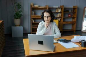 Confident stylish european middle aged senior woman using laptop at home. Stylish older mature 60s lady sitting at table looking at computer screen typing chatting reading writing email. photo