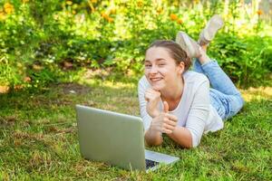 joven mujer acostado en verde césped césped en ciudad parque trabajando en ordenador portátil ordenador personal computadora. Lanza libre negocio concepto foto