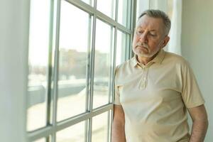 Sad tired lonely disappointed older senior man at home alone. Unhappy grandfather looking sadly outside window experiencing grief relative death bad news. Stressed elder man suffering from loneliness. photo