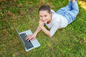 joven mujer acostado en verde césped césped en ciudad parque trabajando en ordenador portátil ordenador personal computadora. Lanza libre negocio concepto foto