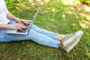 piernas de mujer en césped verde en el parque de la ciudad, manos trabajando en una computadora portátil. concepto de negocio independiente foto