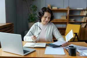 Middle aged senior woman sit with laptop and paper document, Smiling older mature lady reading paper bill pay online at home managing bank finances calculating taxes planning loan debt pension payment photo