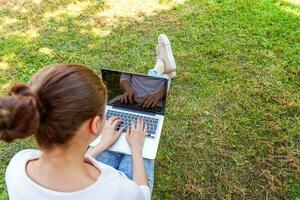 mujer joven sentada en el césped verde en el parque de la ciudad trabajando en una computadora portátil. concepto de negocio independiente foto