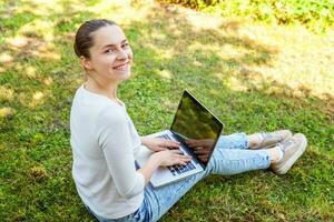 Young woman sitting on green grass lawn in city park working on laptop pc computer. Freelance business concept photo