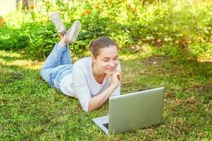 Young woman lying on green grass lawn in city park working on laptop pc computer. Freelance business concept photo