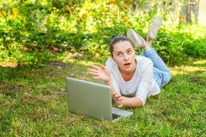 Young woman lying on green grass lawn in city park working on laptop pc computer. Freelance business concept photo