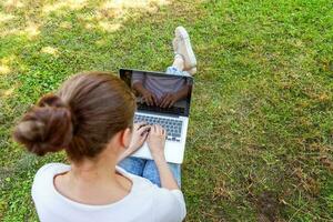 Young woman sitting on green grass lawn in city park working on laptop pc computer. Freelance business concept photo