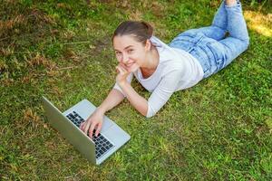 Young woman lying on green grass lawn in city park working on laptop pc computer. Freelance business concept photo