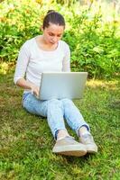 Young woman sitting on green grass lawn in city park working on laptop pc computer. Freelance business concept photo