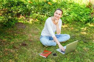 mujer joven sentada en el césped verde en el parque de la ciudad trabajando en una computadora portátil. concepto de negocio independiente foto