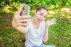 joven gracioso niña tomar selfie desde manos con teléfono sentado en verde césped parque o jardín antecedentes foto