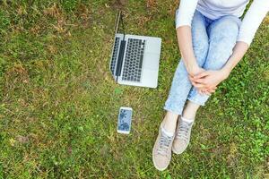 piernas de mujer en césped verde en el parque de la ciudad, manos trabajando en una computadora portátil. concepto de negocio independiente foto