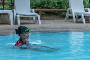 asiático pequeño niña en un traje de baño nadando a el nadando piscina foto