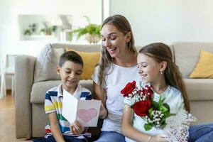 Young mother with a bouquet of roses laughs, hugging her son, and cheerful girl with a card and roses congratulates mom during holiday celebration at home. mothers day photo