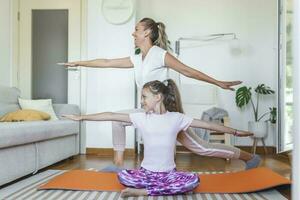 lado ver de positivo joven mujer y pequeño niña en ropa de deporte ejecutando yoga ejercicios mientras formación juntos en moderno ligero Departamento foto