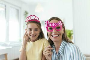 Funny family on a background of bright wall. Mother and her daughter girl with a paper accessories. Mom and child are holding paper crown on stick. photo