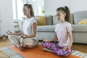 alegre joven mujer y pequeño niña en ropa de deporte hacer ejercicio juntos en moderno ligero habitación. madre y hija practicando yoga foto