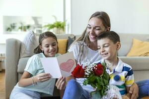 Young mother with a bouquet of roses laughs, hugging her son, and cheerful girl with a card and roses congratulates mom during holiday celebration in kitchen at home photo