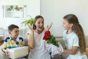 alegre pequeño niña con ramo de flores de rosas flores y el más joven hermano con regalo caja sonriente y felicitando contento mamá en madre día a hogar. contento madres día foto